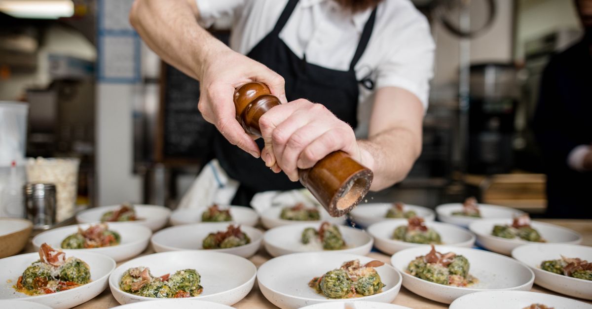 Bearded chef seasoning gourmet dishes with pepper in a busy restaurant kitchen.
