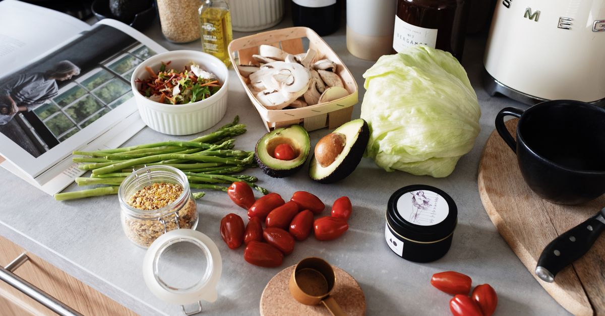From above of various organic vegetables chopped ingredients near cutting board kitchenware appliances and trendy magazine placed on gray countertop in kitchen at home