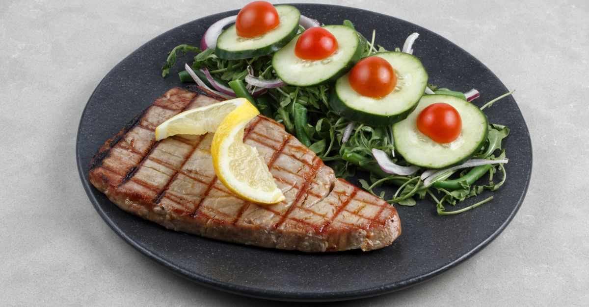 A grilled tuna steak topped with lemon slices served with a fresh cucumber and tomato salad on a dark plate.