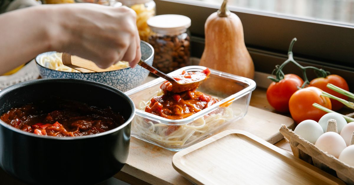 Unrecognizable cook pouring tasty sauce from saucepan into container with pasta while cooking at counter with ingredients in light kitchen