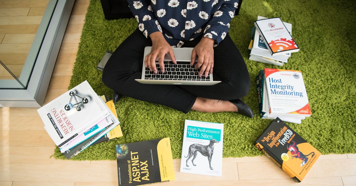 Woman sitting on green rug working on laptop, surrounded by technology books in a modern room.