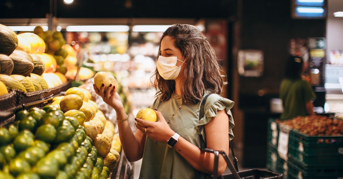 A woman wearing a mask selects fresh fruit in a grocery store aisle.