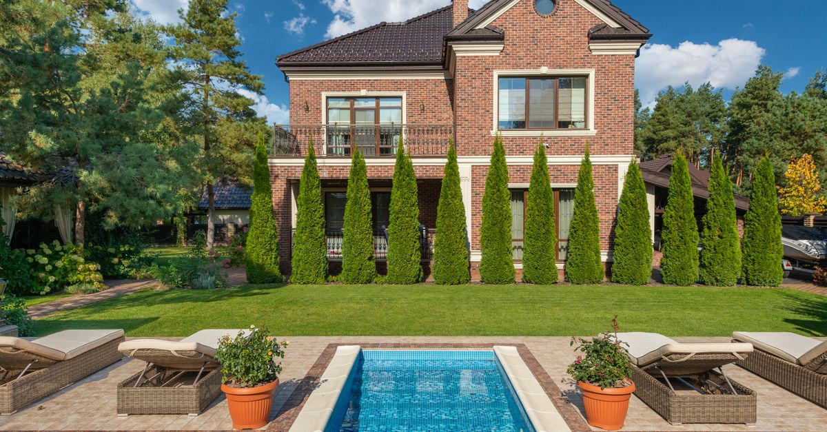 Facade of brick dwell villa with green bushes on lawn and sunbeds near swimming pool