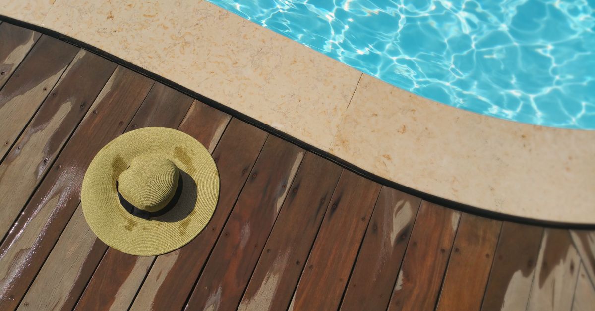 A straw hat rests on a wooden deck beside a sparkling blue swimming pool, perfect for summer relaxation.