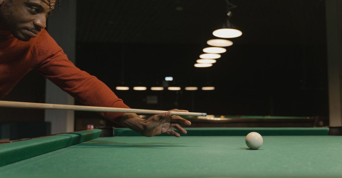 An adult man aiming a cue stick at a cue ball on a billiards table indoors.