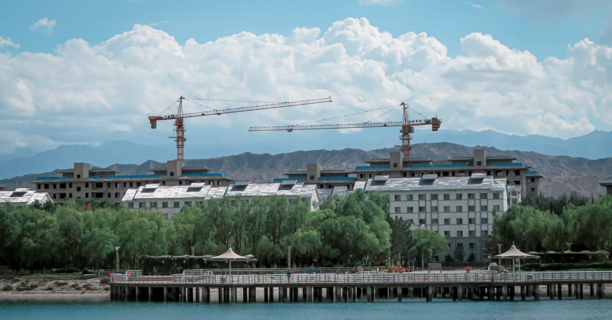 Urban construction site with cranes, buildings by water, mountains in the background.