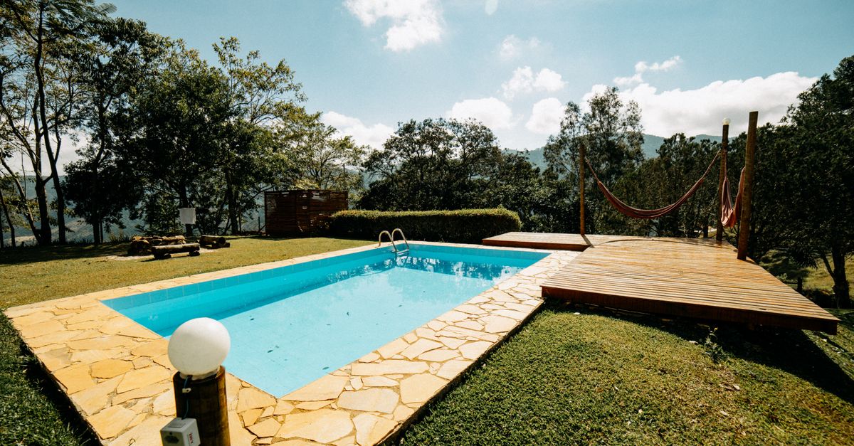 A serene outdoor pool with a wooden deck and hammock surrounded by lush greenery on a sunny day.