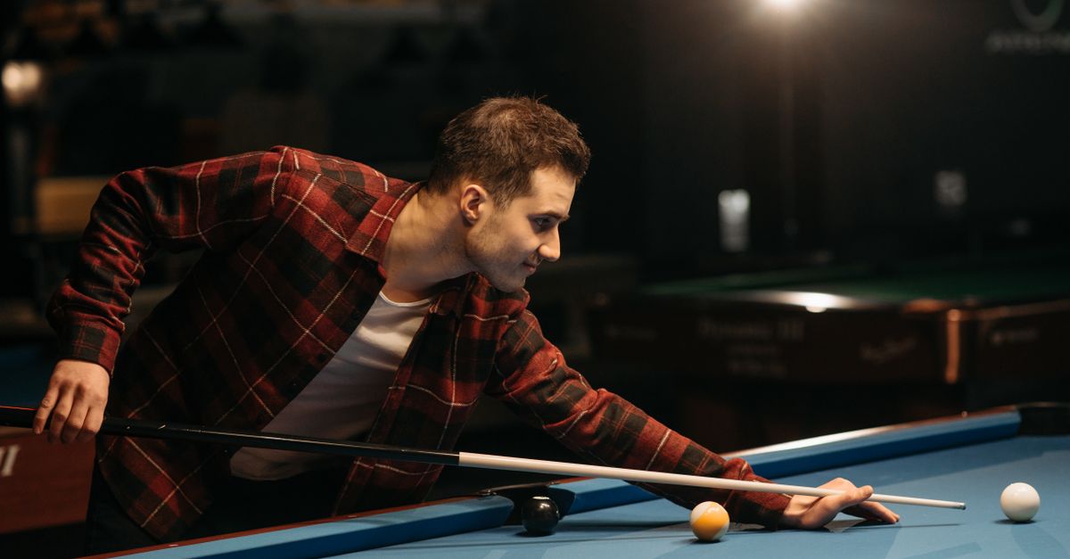 Man in plaid shirt aiming a shot on a billiard table indoors.