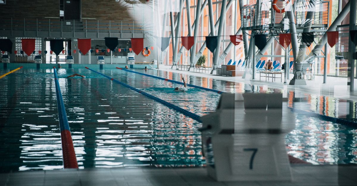 Through glass wall view of starting platform with number against textile garland above shiny pool with unrecognizable sportspeople swimming during training