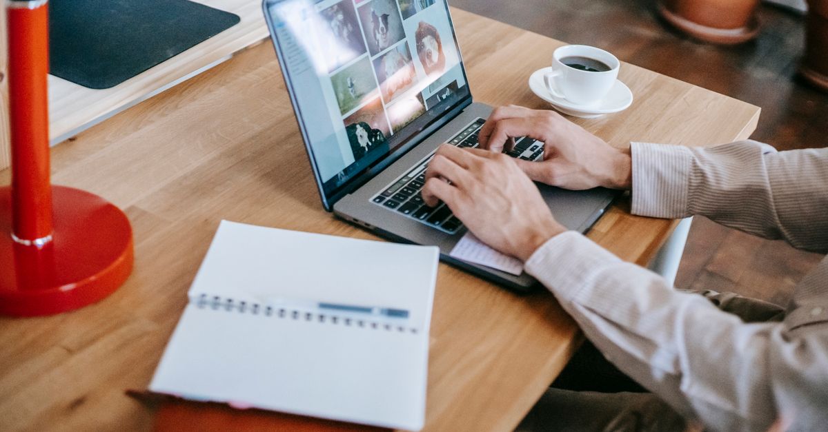 High angle of crop faceless male remote worker in casual clothes sitting at wooden table while using laptop near notepad and drinking coffee in light room