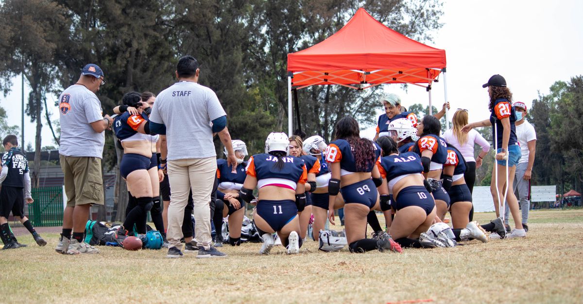 A women's flag football team huddles under a red canopy on a field, preparing for the game.