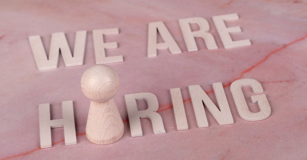 Wooden letters spelling 'We Are Hiring' on a pink marble surface with a pawn piece.