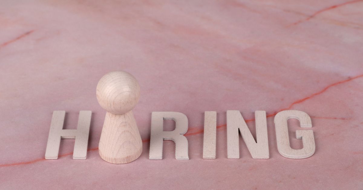 Wooden letters spelling 'hiring' on pink marble background with a peg.