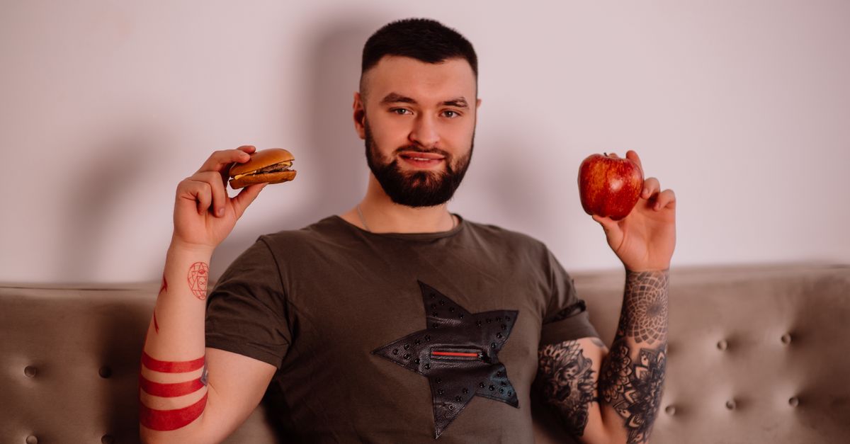 Bearded man pondering diet choice with apple and burger. Indoors with tattoos visible.