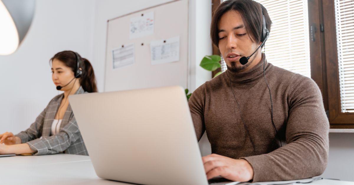 Two call center employees working together, wearing headsets, in an office setting.