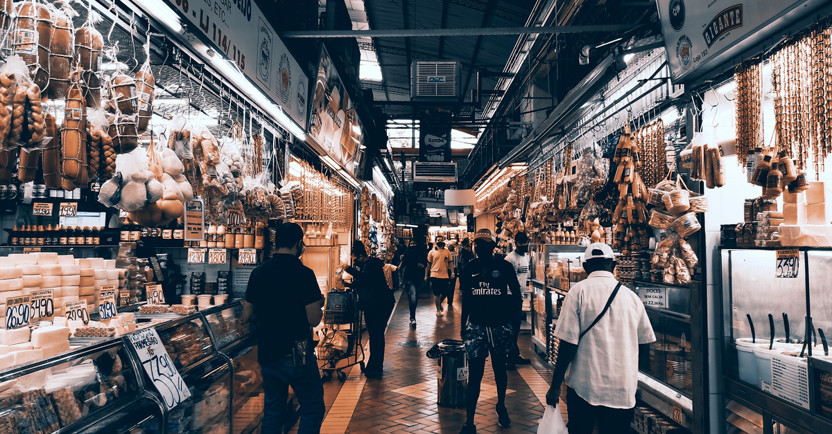 A bustling indoor market aisle filled with diverse shops and people shopping.