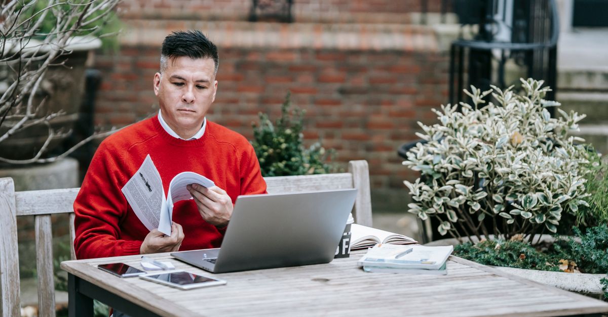 Adult male reading outside with a laptop on a wooden table, embracing remote work lifestyle.