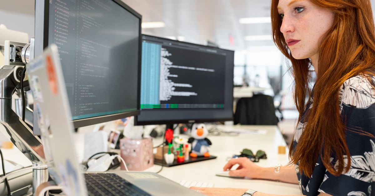 A focused female software engineer coding on dual monitors in a modern office.