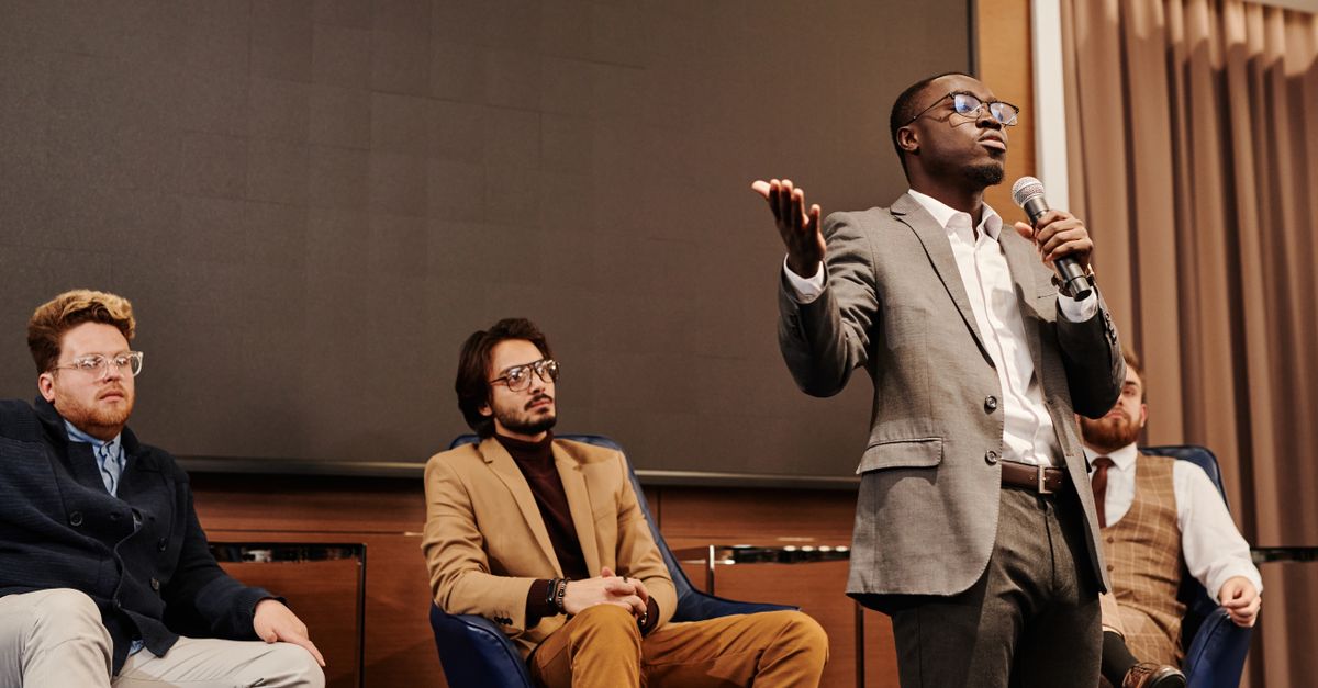 A diverse group of professionals engaged in a business panel discussion with a speaker presenting.