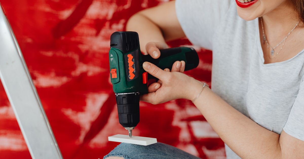 A smiling woman using a cordless drill, engaged in a home renovation project with a red background.