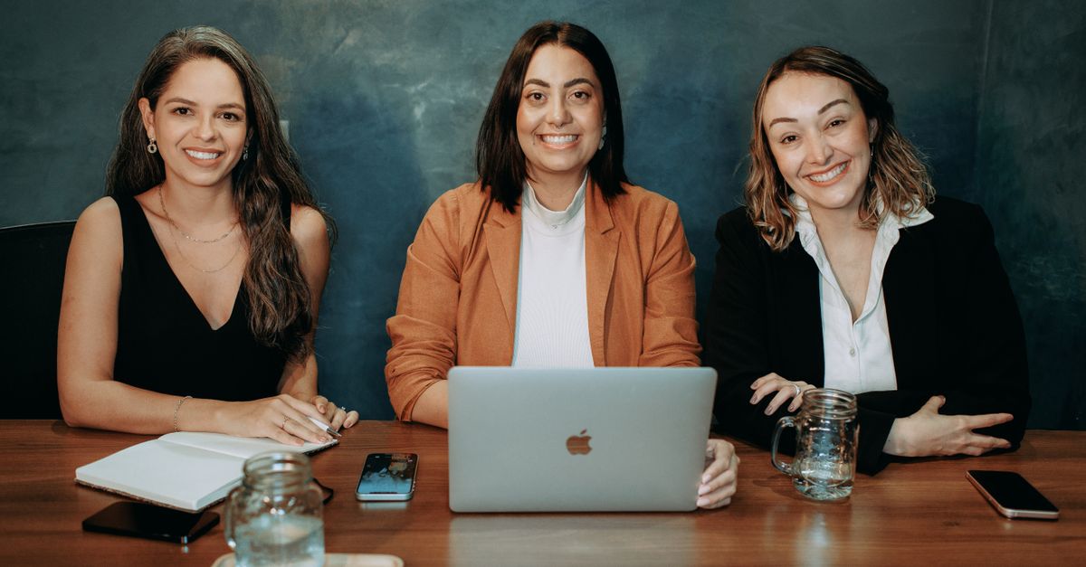 Three professional women sitting at a wooden desk in an office with laptops and notebooks.