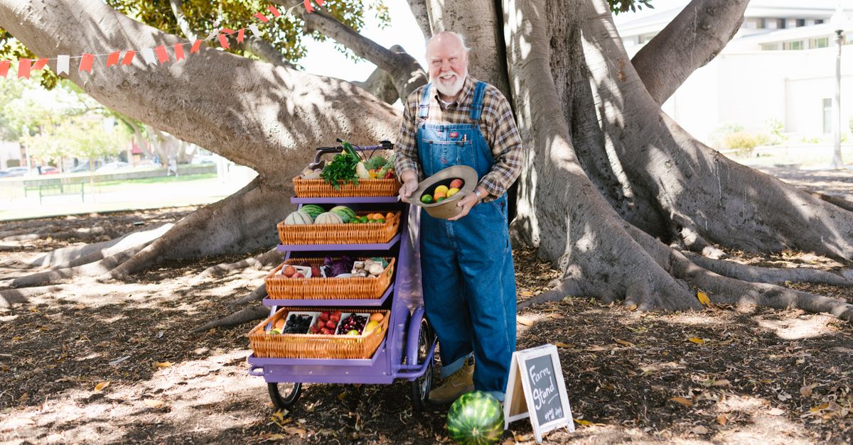 Elderly farmer smiling by his outdoor produce stand with fresh fruits.