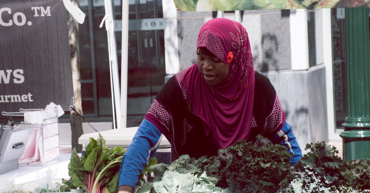 Woman in headscarf selling fresh greens at outdoor market stall.