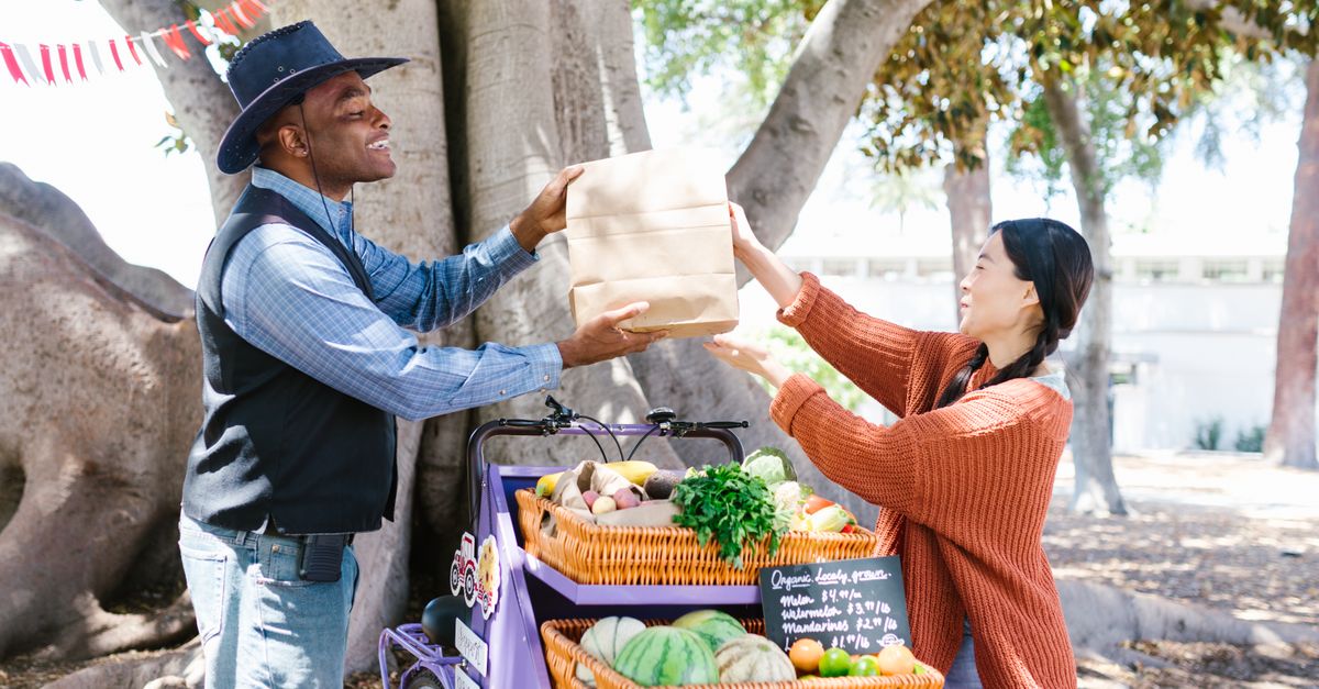 A vendor hands a paper bag to a customer at an outdoor farmer's market filled with fresh produce.