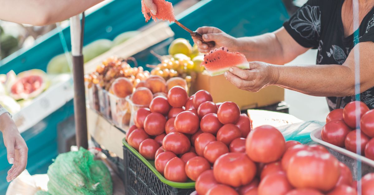 A vendor hands a watermelon slice at a vibrant outdoor market with various fruits.