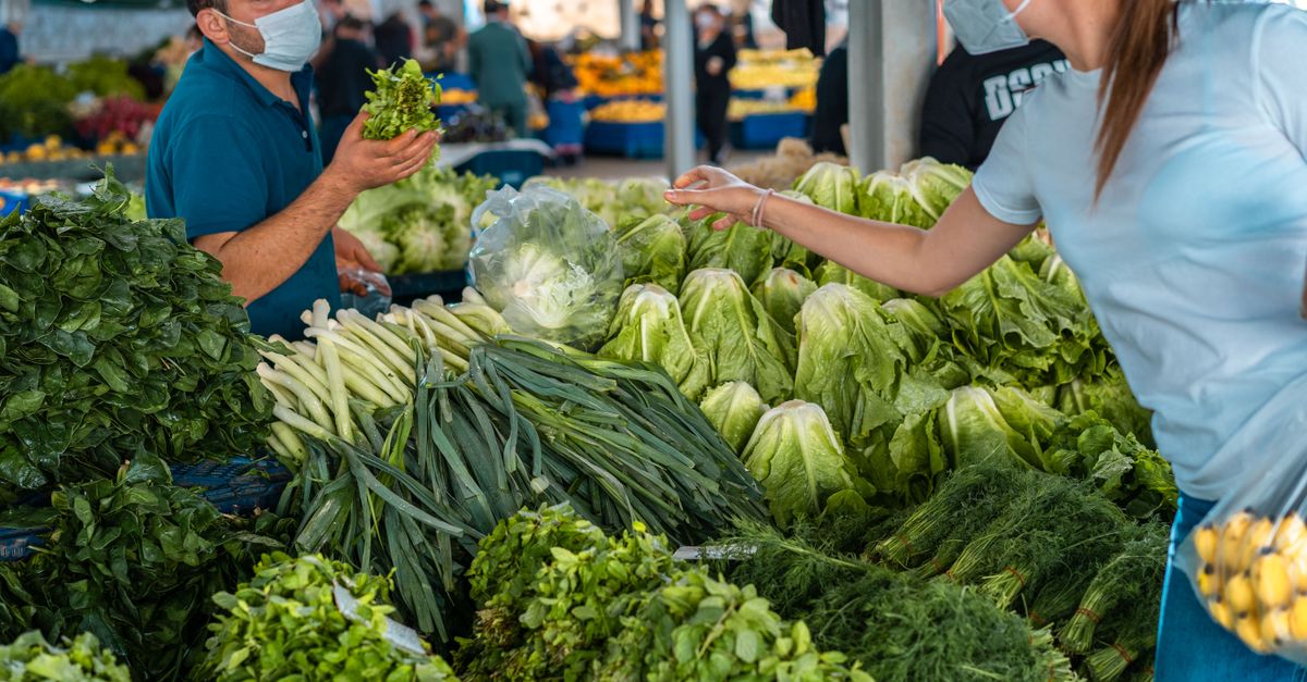 A merchant selling fresh vegetables to a masked woman at an outdoor market.