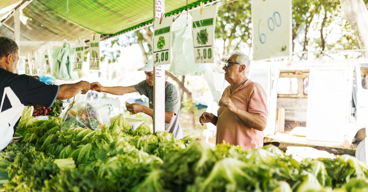 Customers purchasing fresh vegetables at an outdoor farmers market under green canopy.