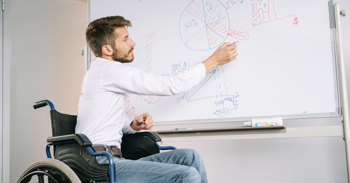 Man in a wheelchair giving a business presentation in an office using a whiteboard with graphs and charts.
