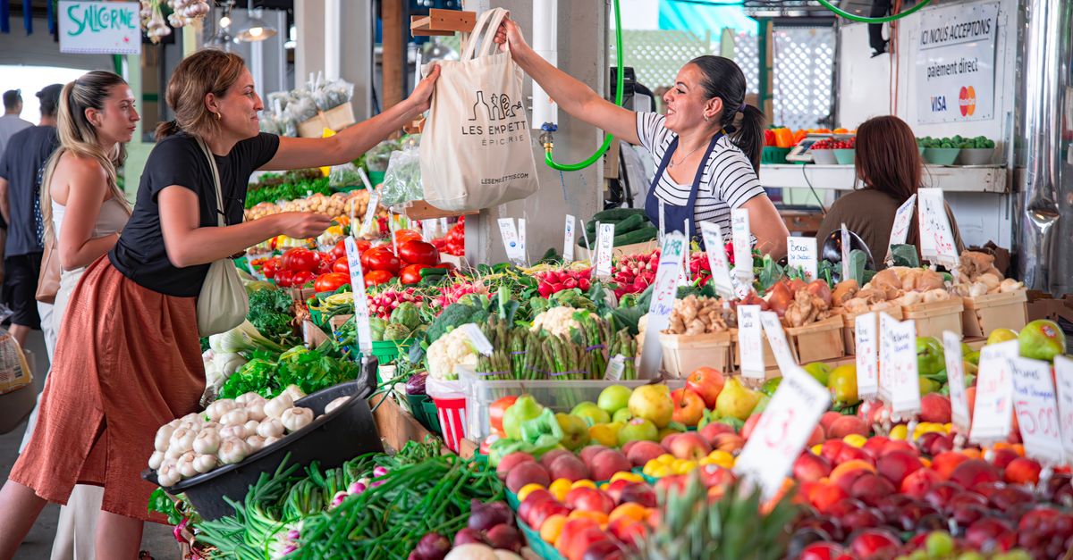 Women shopping at a colorful outdoor market stall filled with fresh fruits and vegetables. Smiles and interaction.