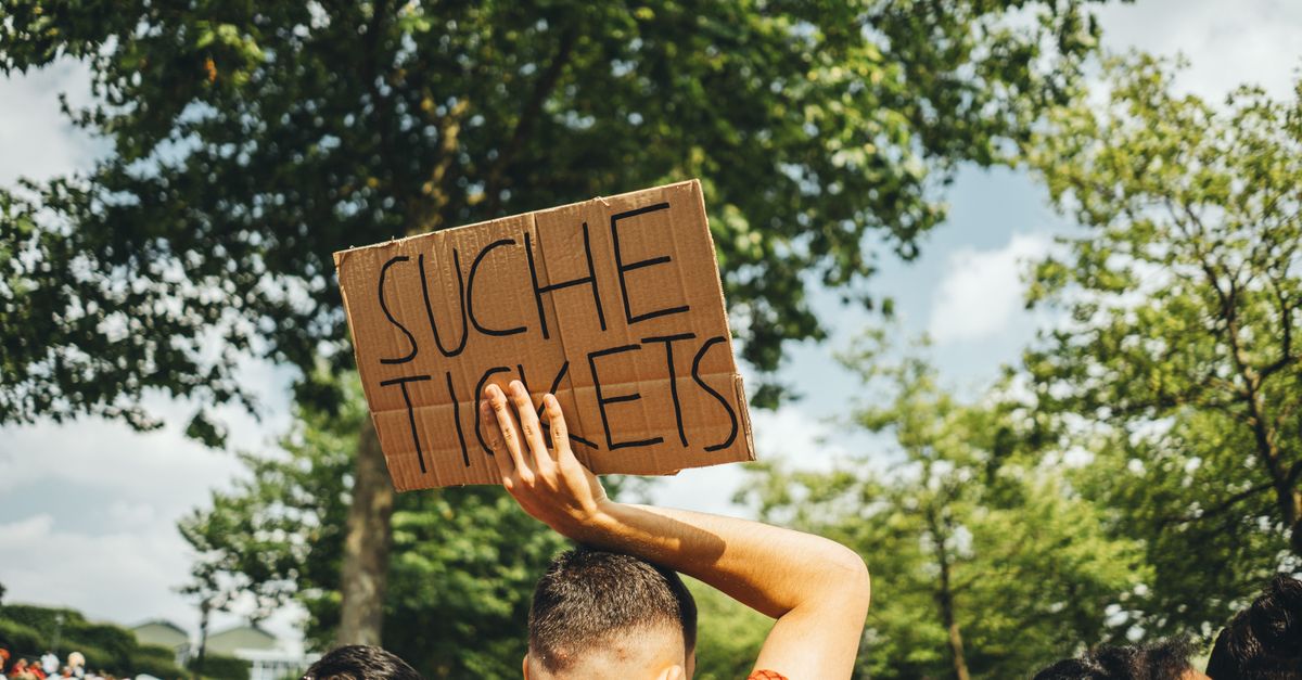 A man in a crowd holds a sign reading 'Suche Tickets' at an outdoor event, seeking extra admissions.