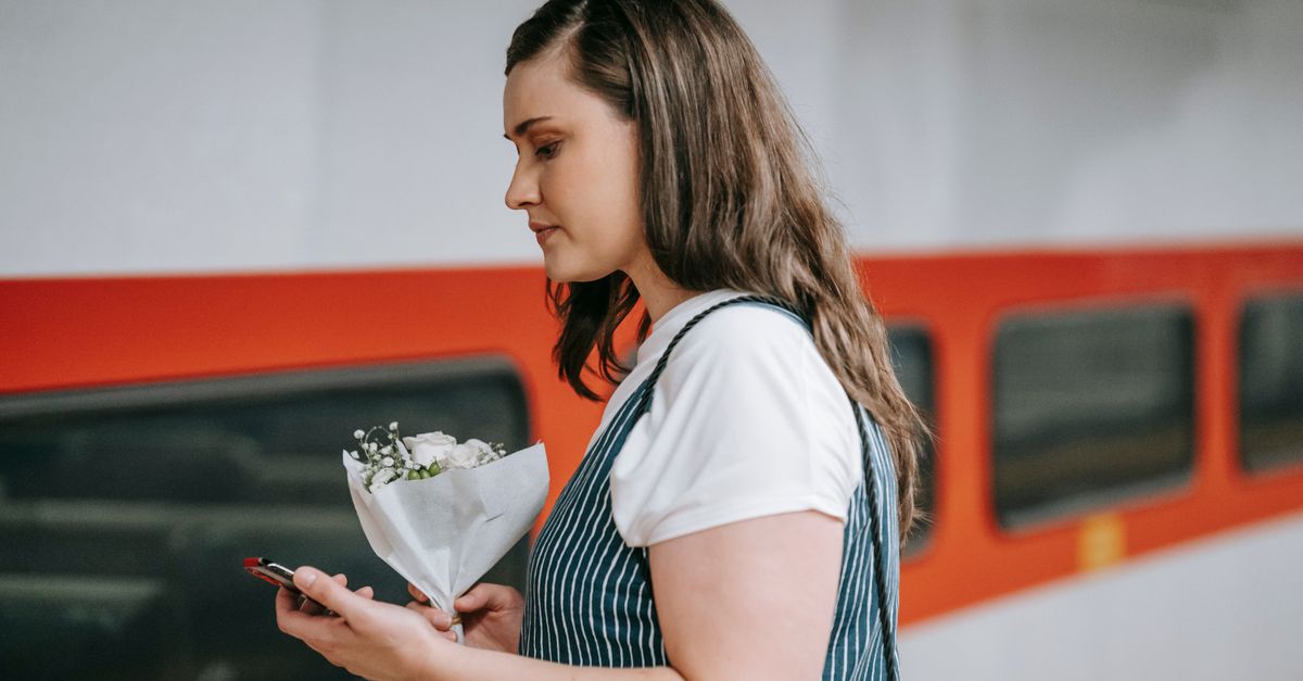 A woman stands at a train station holding flowers and using her phone.