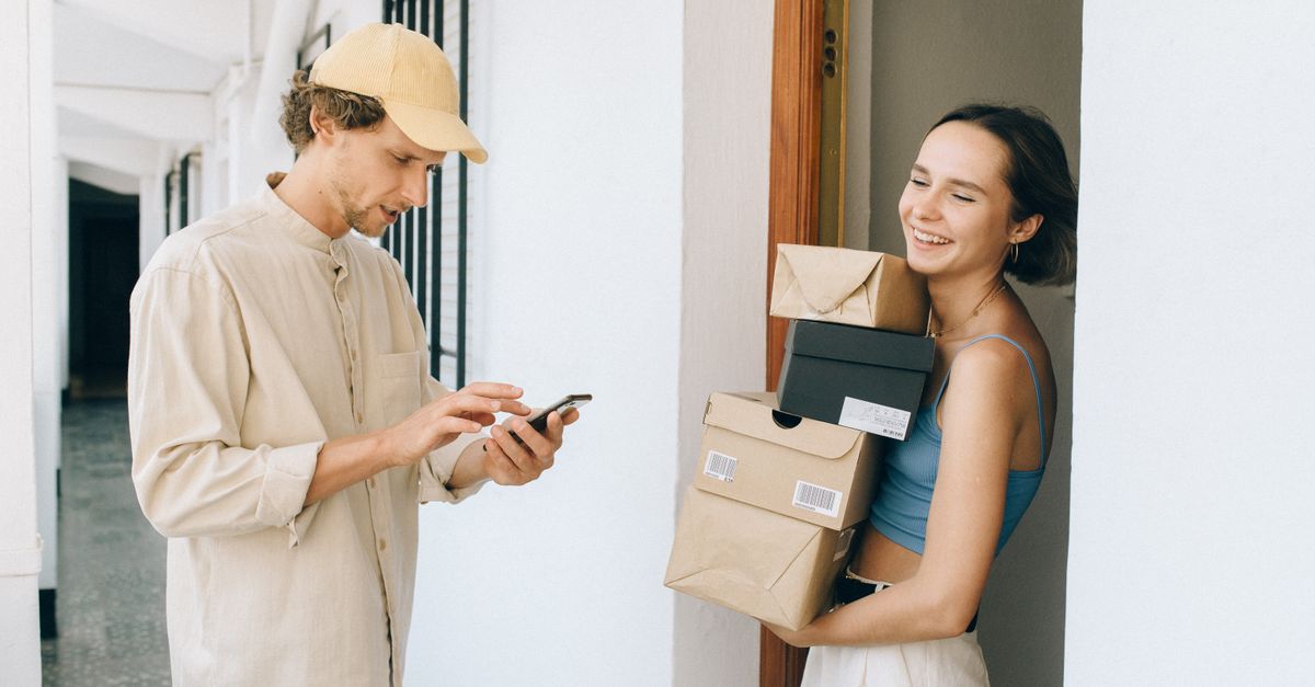 A cheerful woman receives a delivery from a person in a hallway, using a smartphone for online payment.
