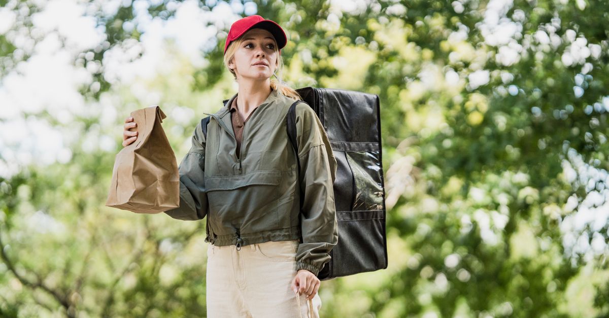 A woman in a red cap delivers meals in a park, holding paper bags.