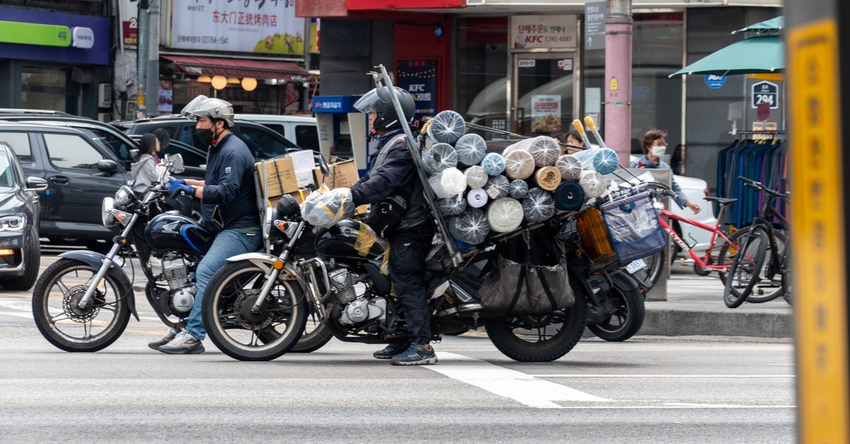 Motorcyclists carrying large cargo loads wait at a city intersection near recognizable fast-food outlets.