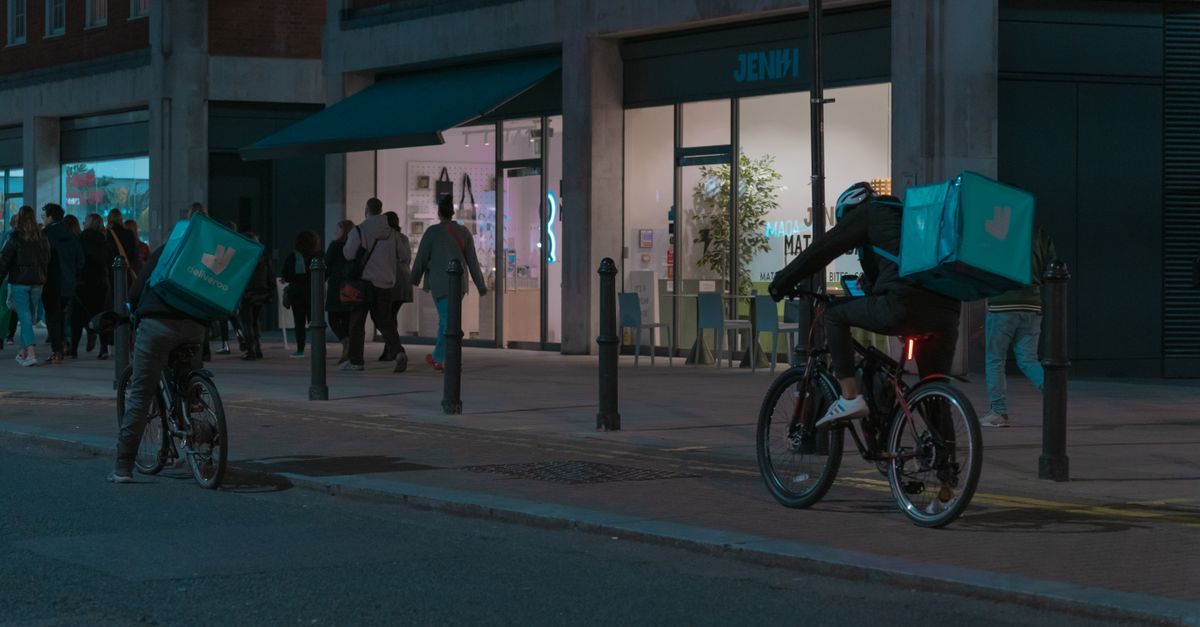 Bicycle delivery workers on city street at night, near a modern storefront with pedestrians.