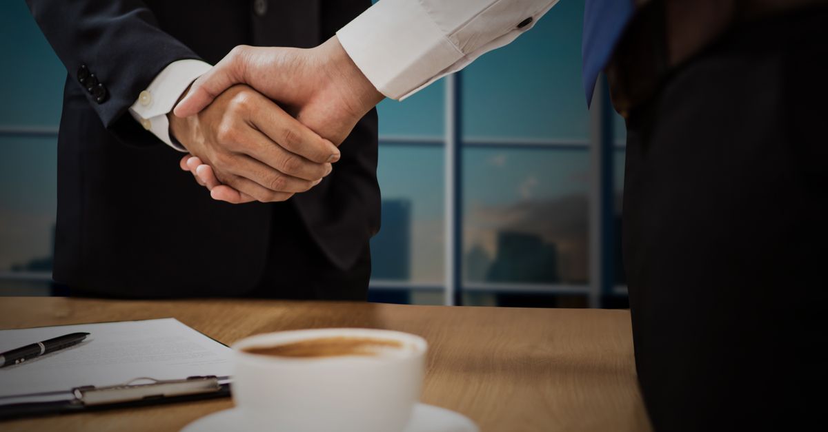 Two professionals shaking hands across a desk with cityscape in the background.