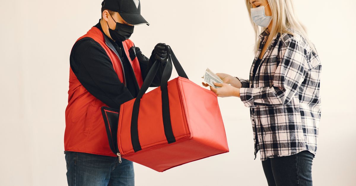 Young blond woman in checkered shirt and medical mask paying cash to young delivery man in protective mask and gloves during safety food delivery in studio