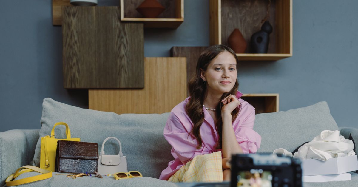 Brunette woman vlogging on sofa with handbags and accessories in modern interior.