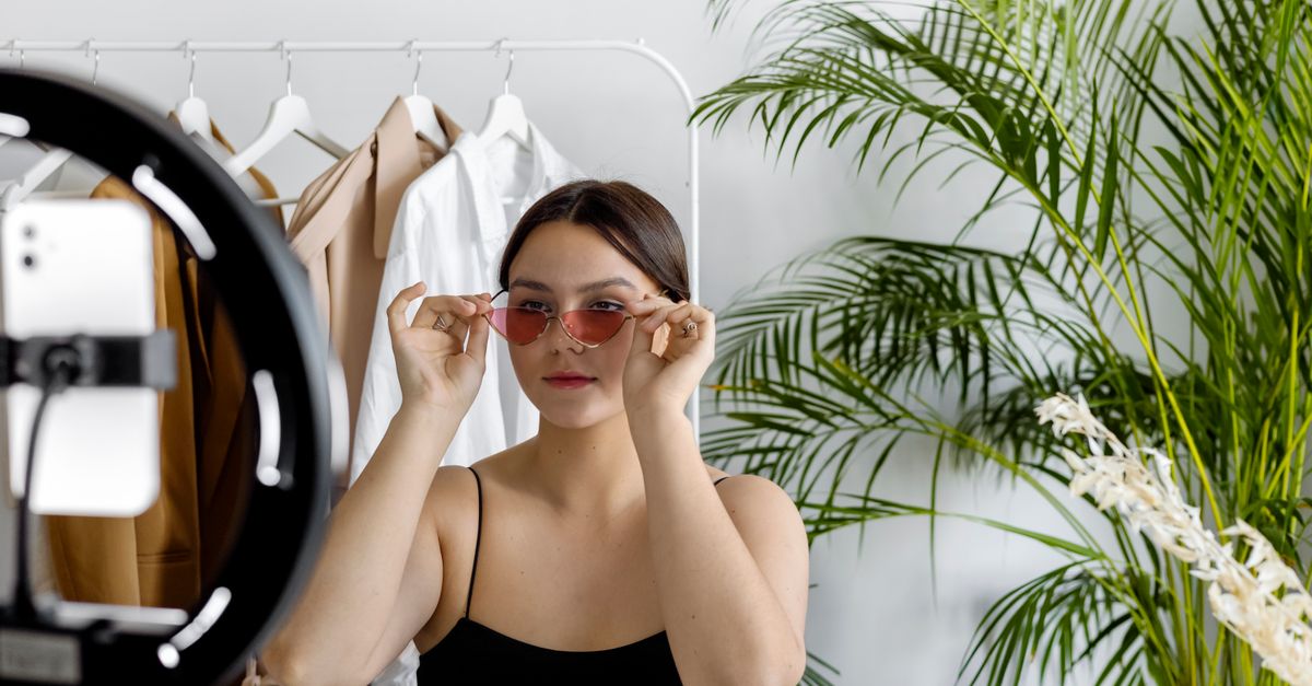 Stylish woman wearing sunglasses recording fashion content with a ring light indoors.