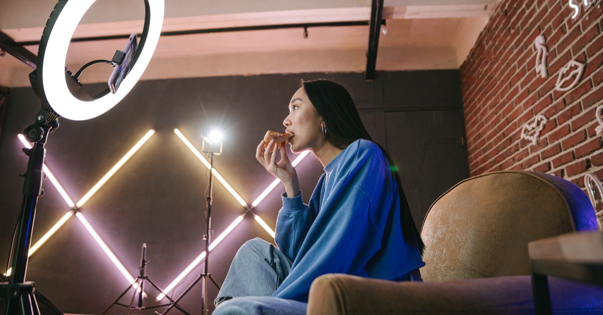 Asian woman content creator eating pizza while recording a video indoors with ring light.