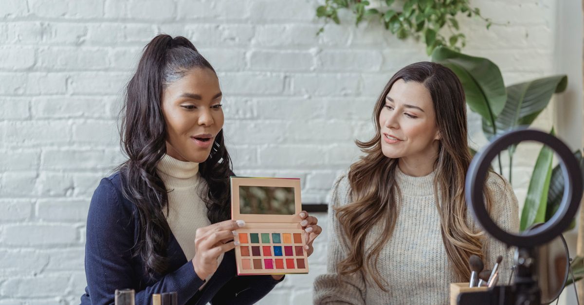 Young African American female blogger demonstrating palette of colorful eyeshadow while recording vlog review on smartphone placed on tripod with friend
