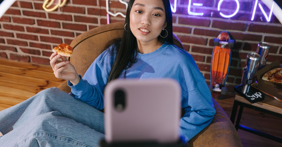Young Asian woman streaming video while enjoying pizza in a cozy setup with neon lights.