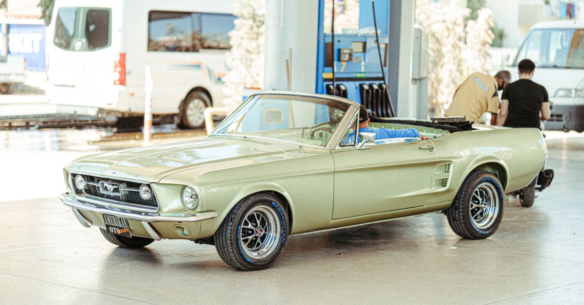 Classic Ford Mustang convertible parked at a Turkish gas station with people around.