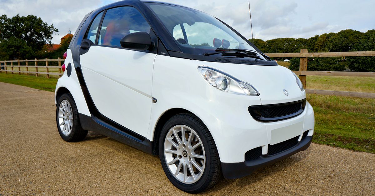 A white Smart Fortwo car parked outdoors with clear sky and greenery.