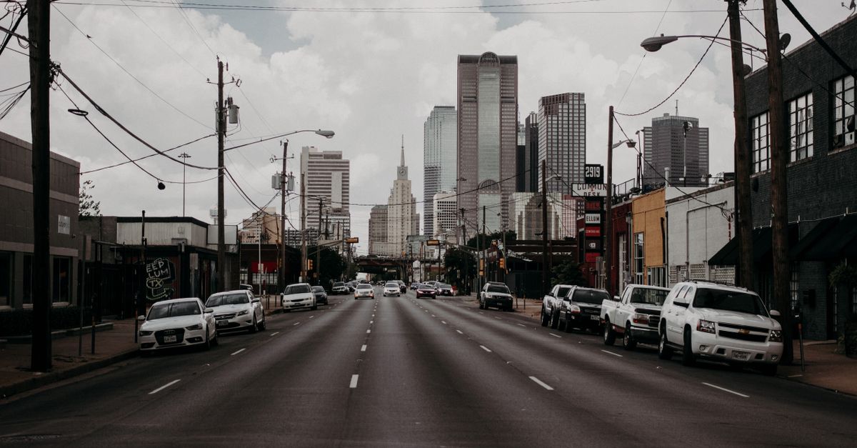 A city street leads to towering skyscrapers under an overcast sky, featuring cars and urban infrastructure.