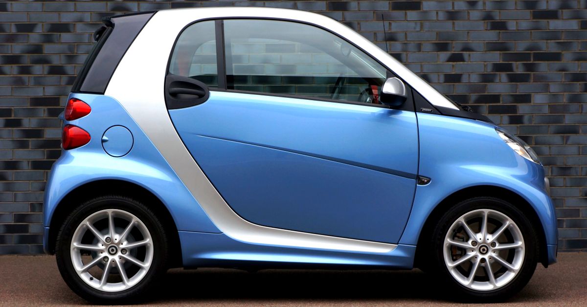 Side view of a modern blue smart car against a brick wall, showcasing compact design.